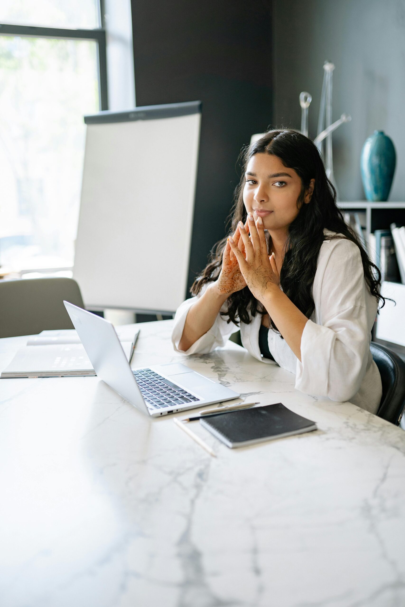 A focused businesswoman in a white shirt working at a modern office desk with a laptop and notepad.